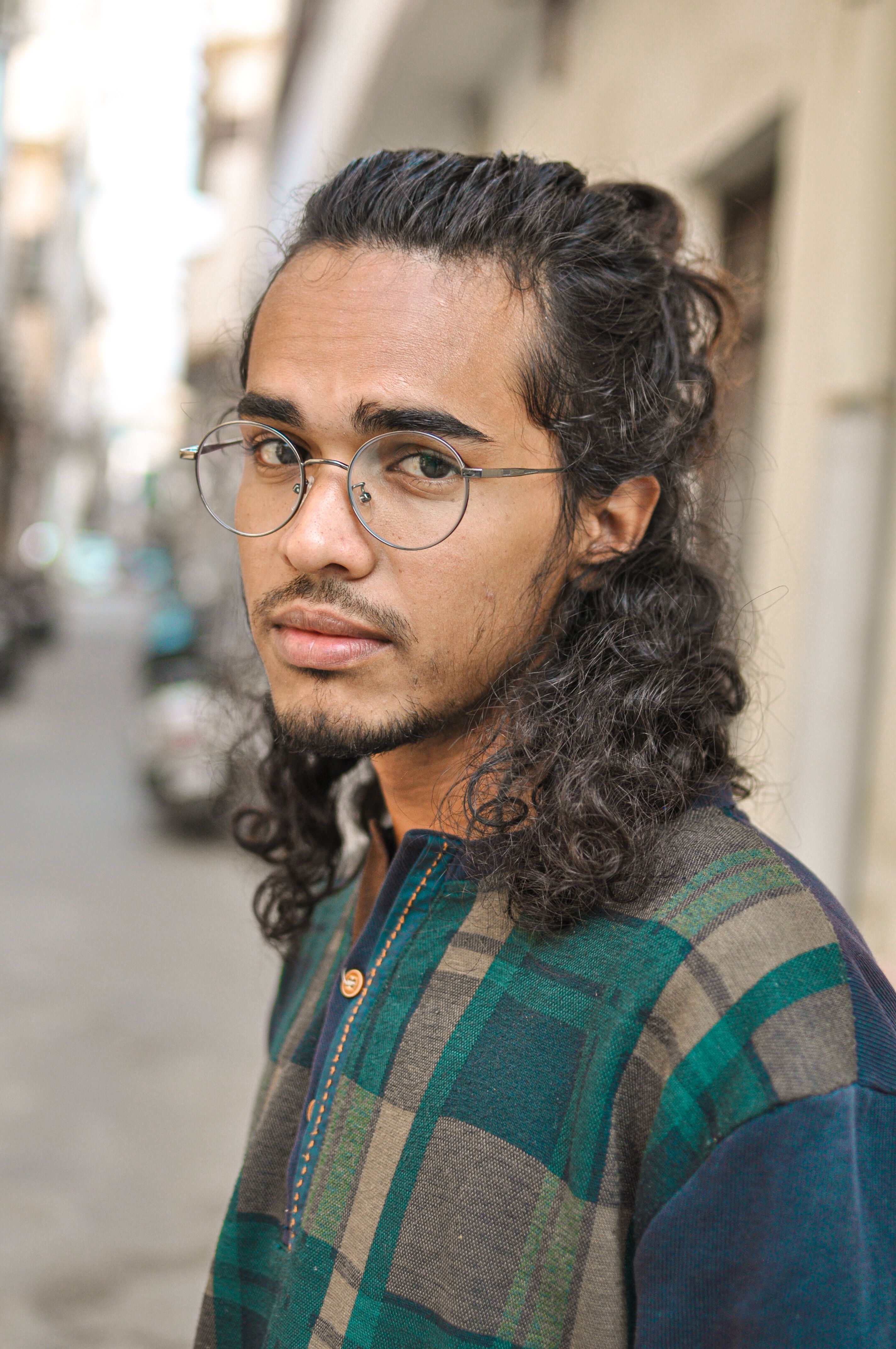 A serene portrait of a young man with curly hair and glasses in a bustling street of Ludhiana, India.
