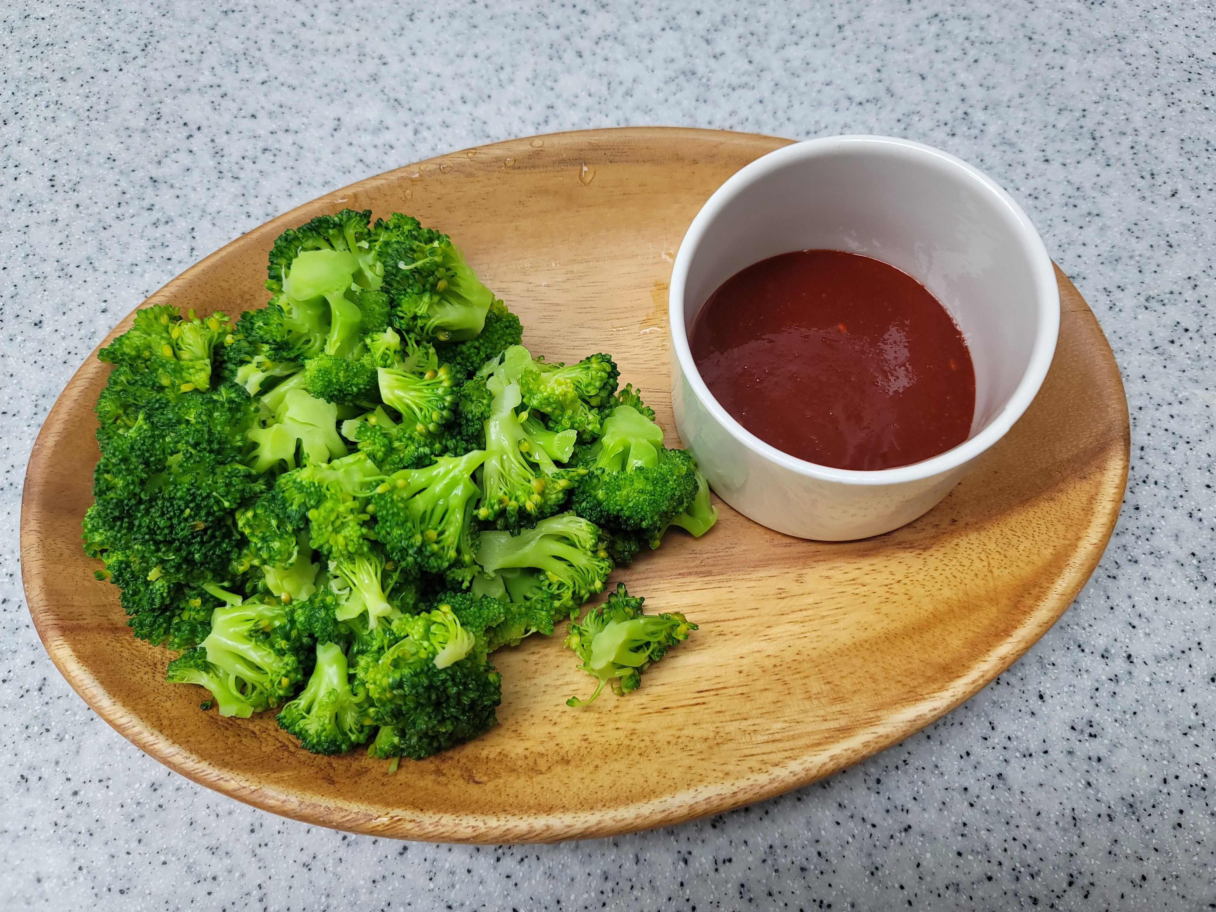 A wooden plate with fresh steamed broccoli and a side of rich tomato sauce.
