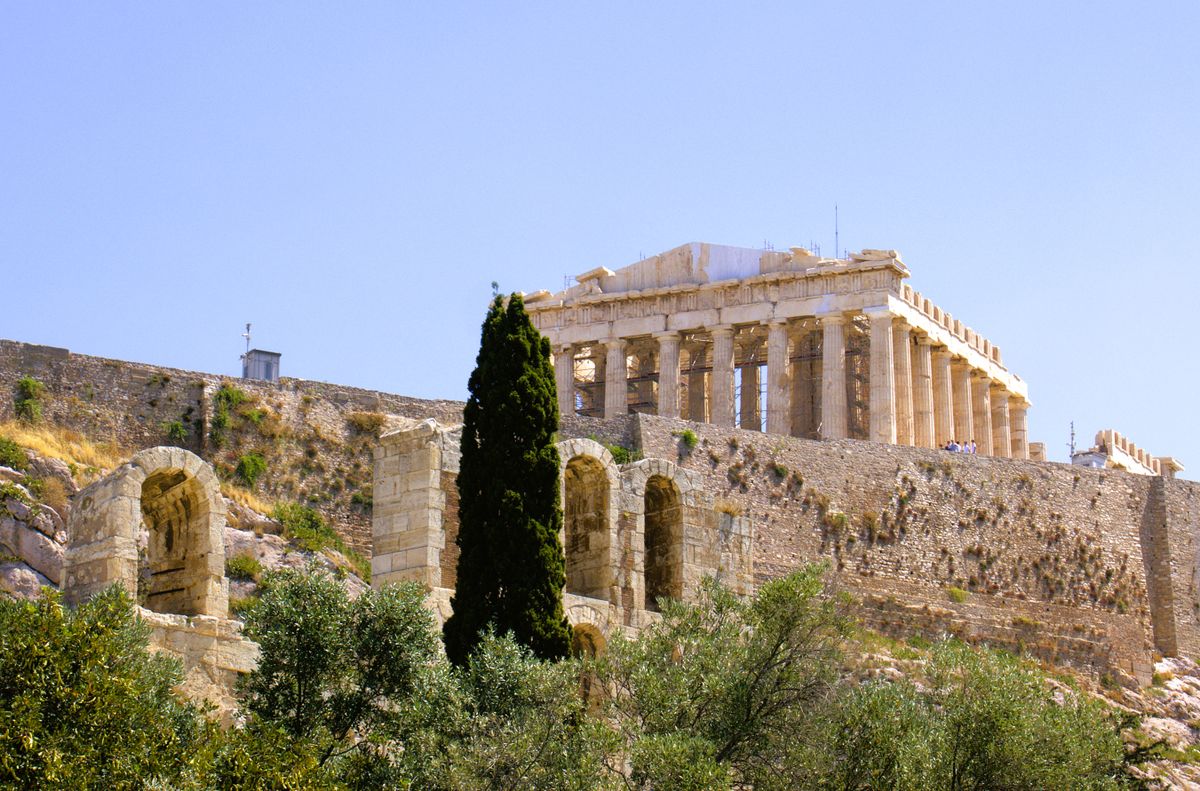 The Parthenon seen from below the fortified walls of the Acropolis in Athens, framed by cypress and olive trees