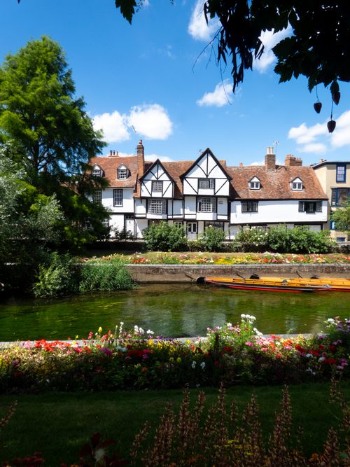 Free Picturesque Tudor-style cottage with garden by a tranquil river under a clear blue sky. Stock Photo