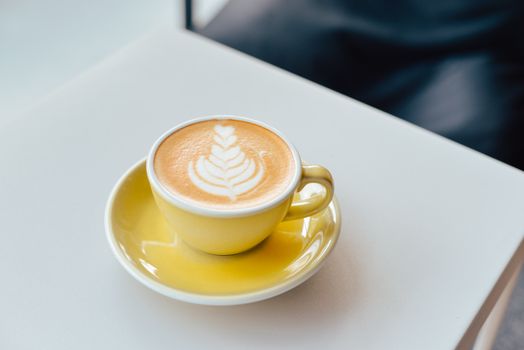 Close-up of a latte with artistic foam design in a yellow cup on a white table. Bright and inviting setting.