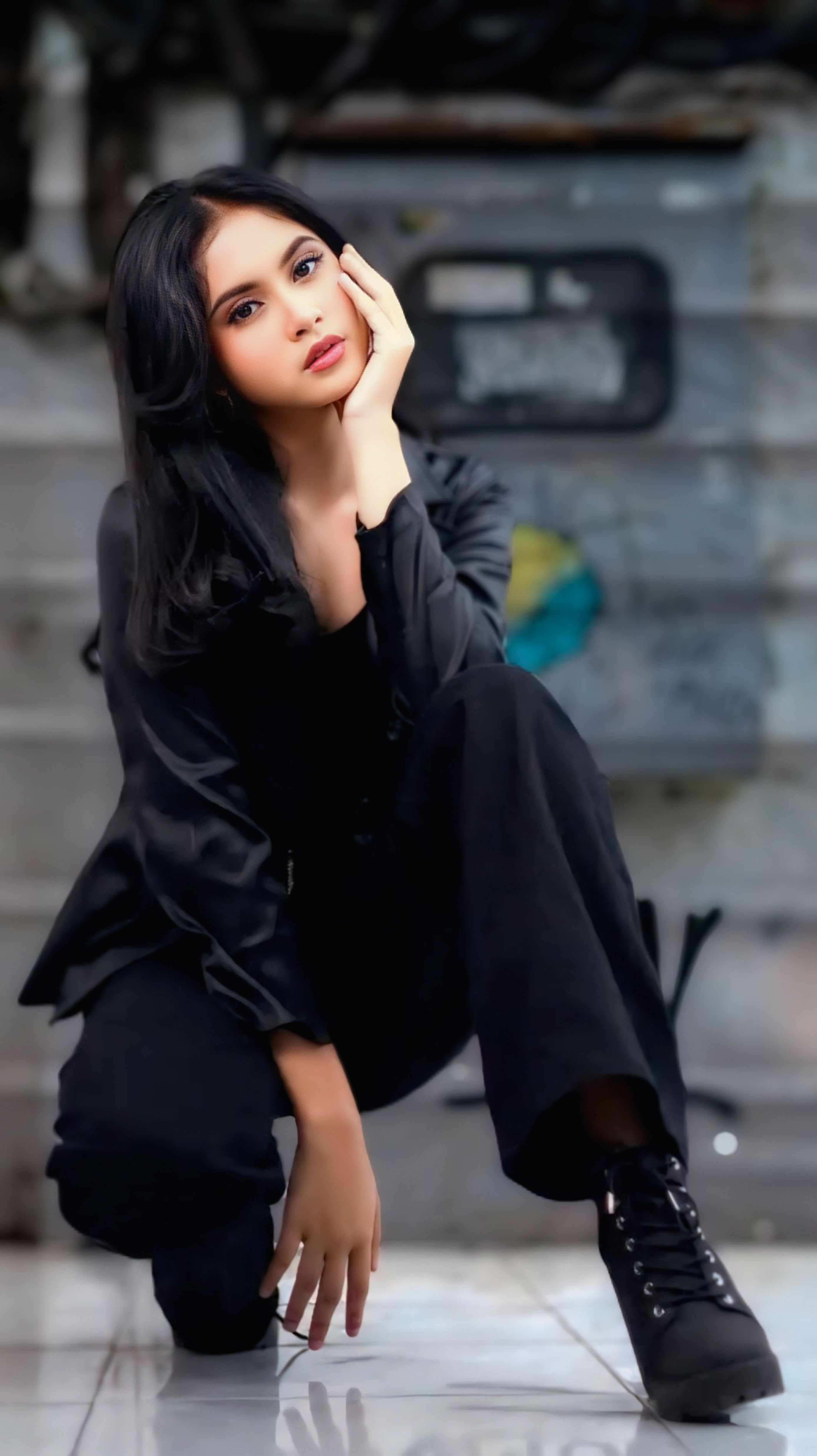 Fashionable young woman in black attire posing outdoors with an urban backdrop.