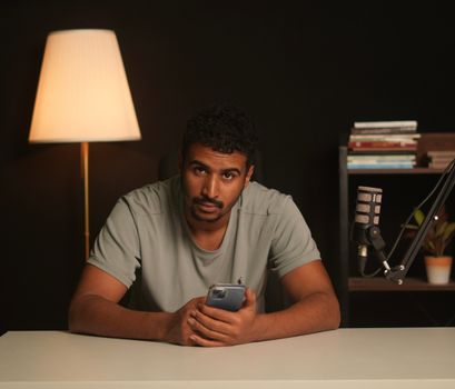 A man in a podcast studio holding a smartphone, with a microphone and books in the background.
