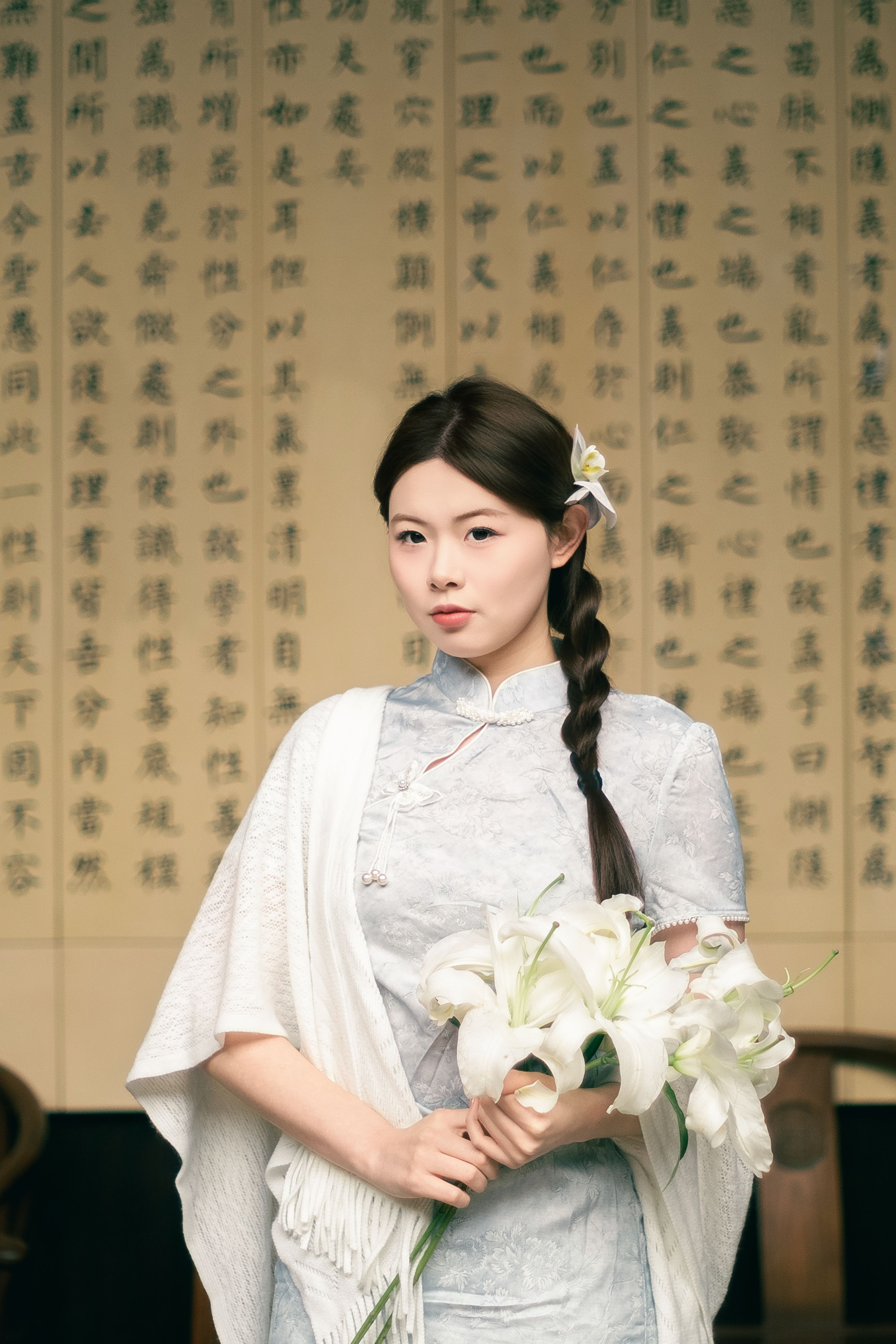 A woman in traditional attire holding lilies, standing before an ornate background with text.