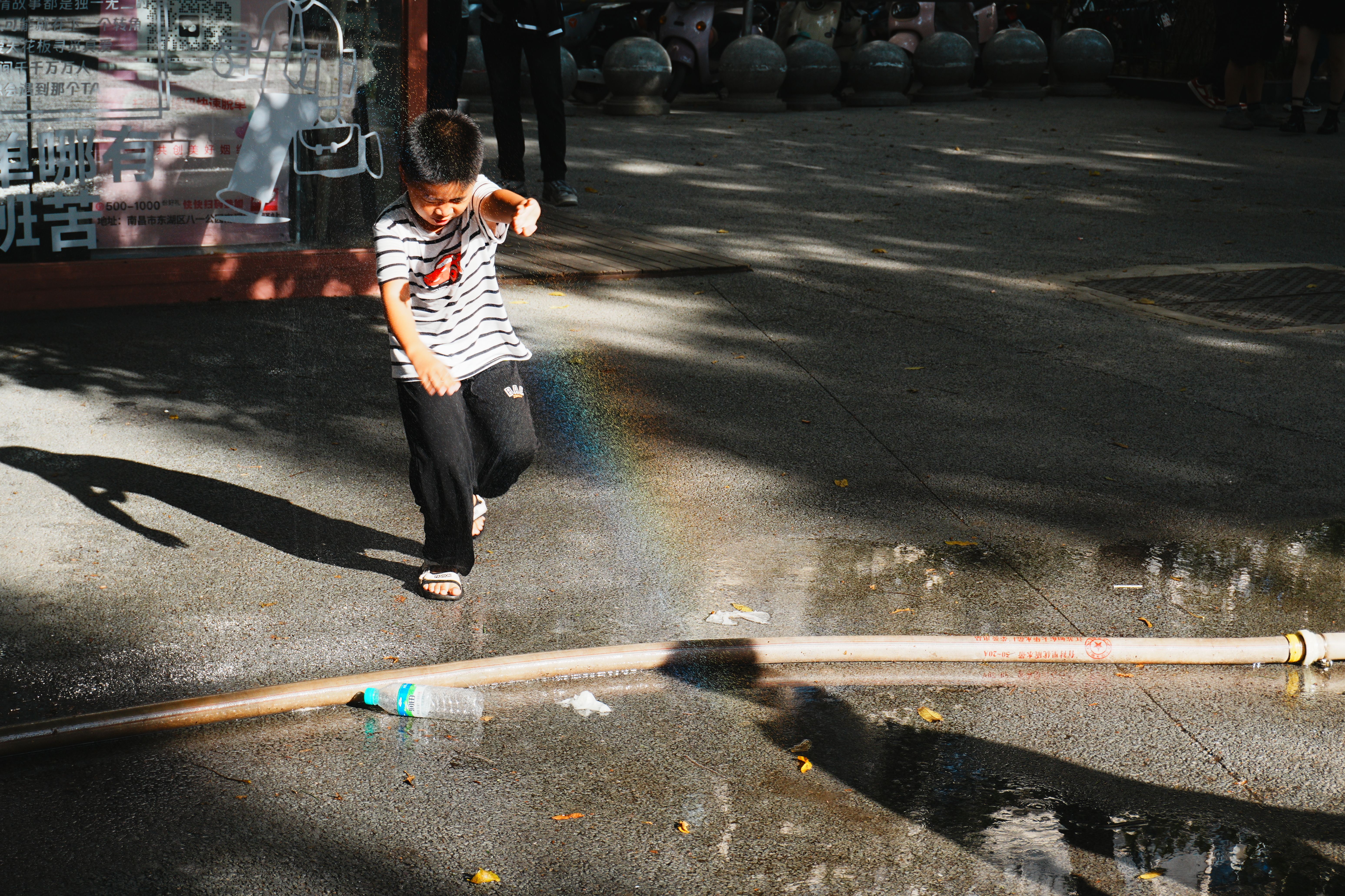 A joyful child splashes water in a sunny street, creating a rainbow reflection.