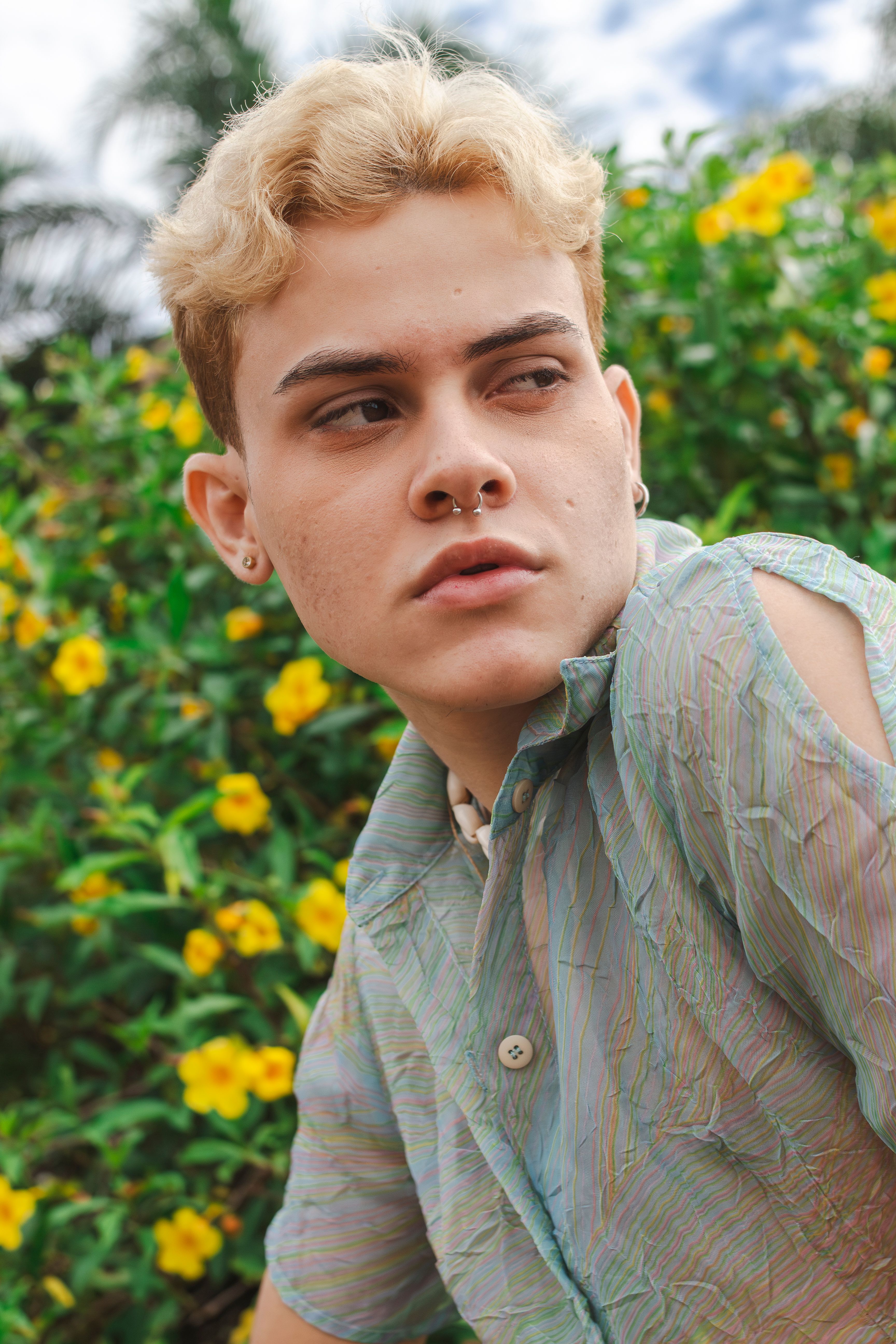 Young adult with piercings poses confidently among yellow flowers in an outdoor garden.