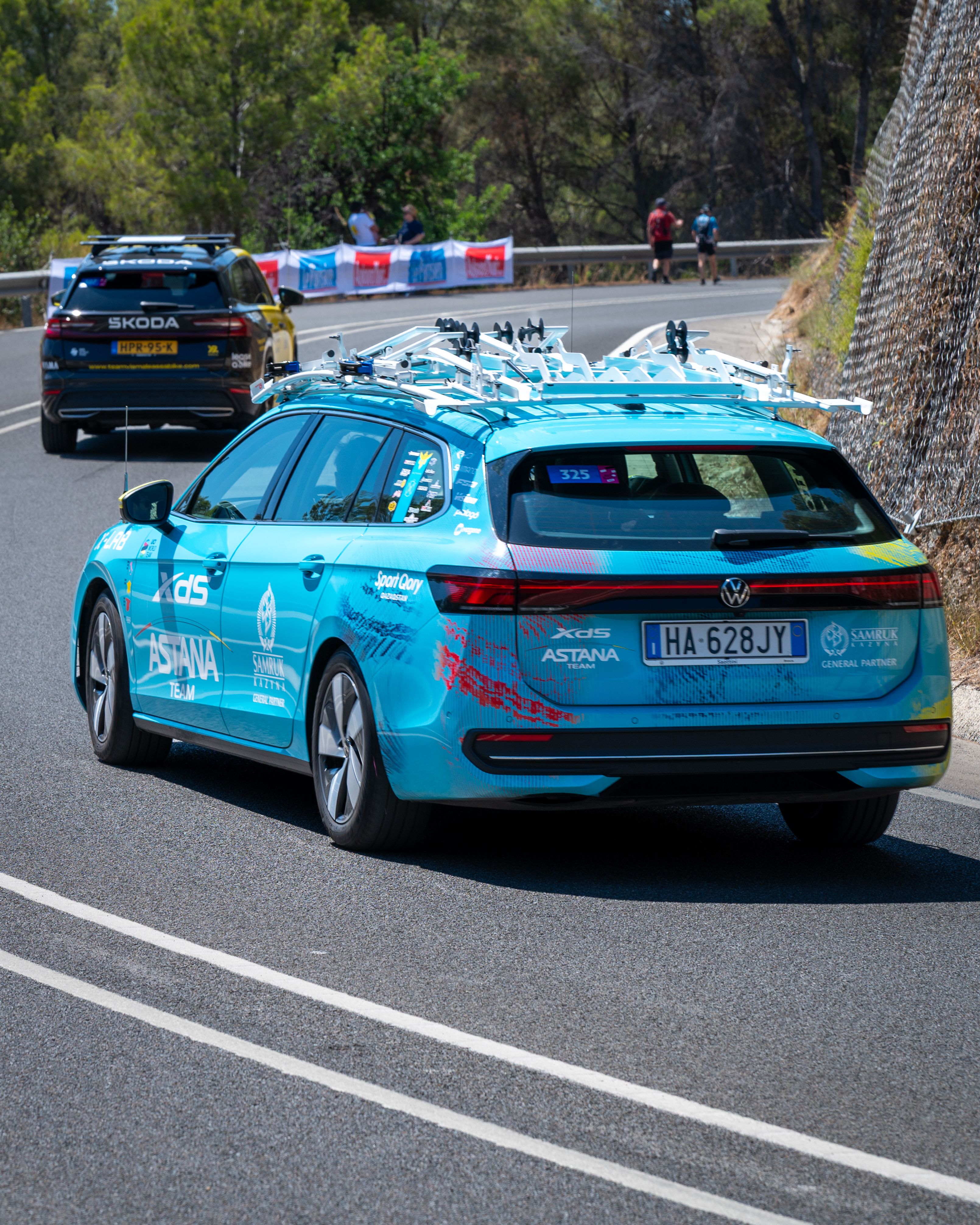 Support vehicles for cycling race drive through a scenic forest road, ensuring athletes' safety and logistics.