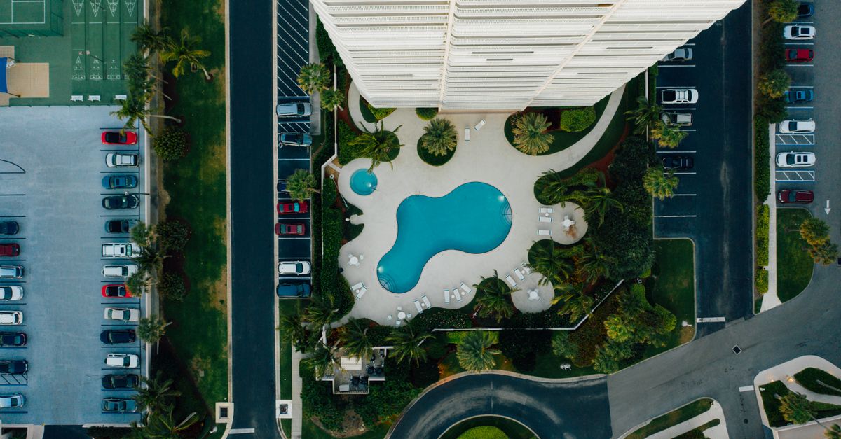 High-angle view of a swimming pool beside a tall urban building with surrounding parking lots.