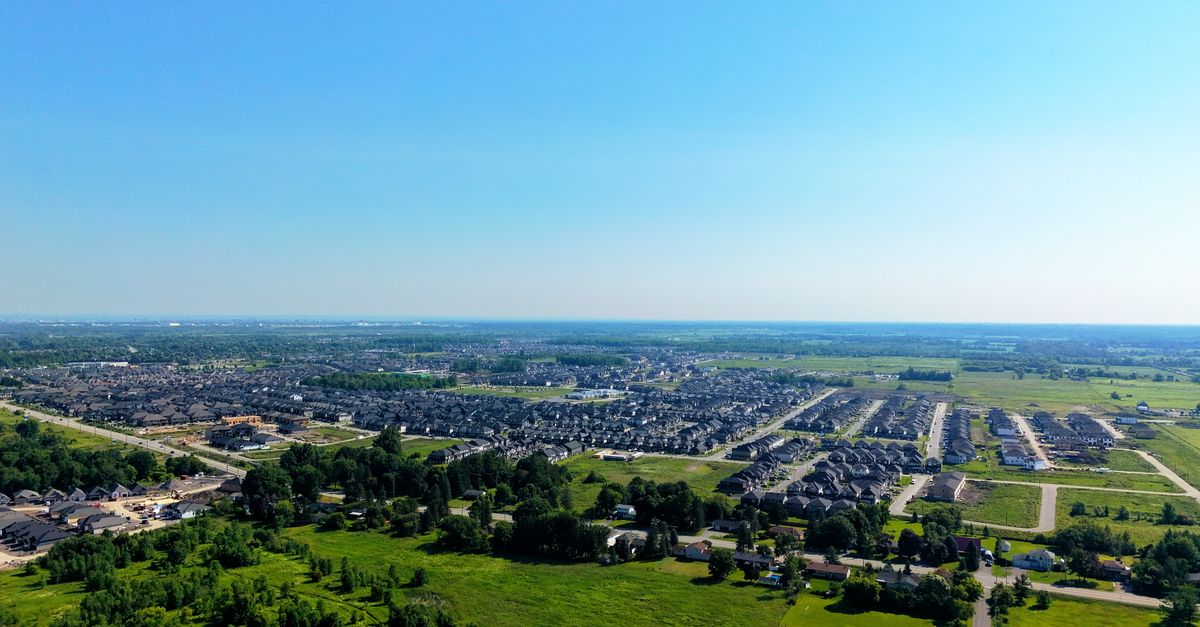 Aerial shot of a residential suburb surrounded by green fields under a blue sky.