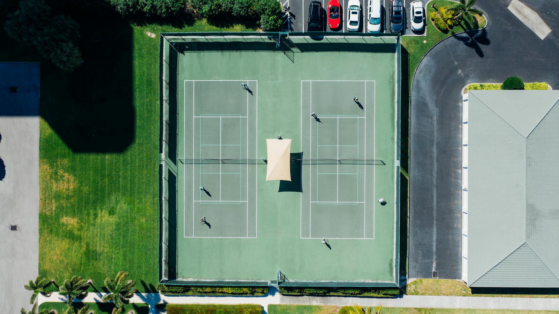 Drone shot capturing a tennis match on dual courts with players and parked cars under clear summer skies.