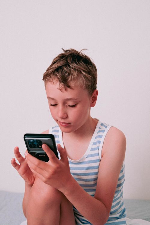 Free A young boy in a striped shirt focuses on his smartphone indoors. Stock Photo