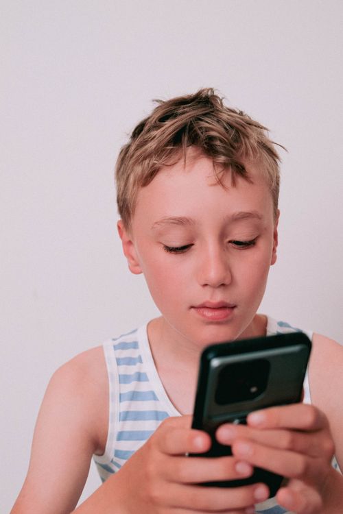 Free A young boy in a striped shirt focuses intently on a smartphone, sitting indoors. Stock Photo