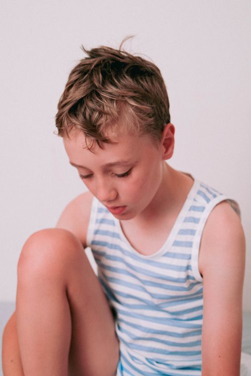 Free Young boy in a striped tank top, seated indoors, looking down thoughtfully. Stock Photo