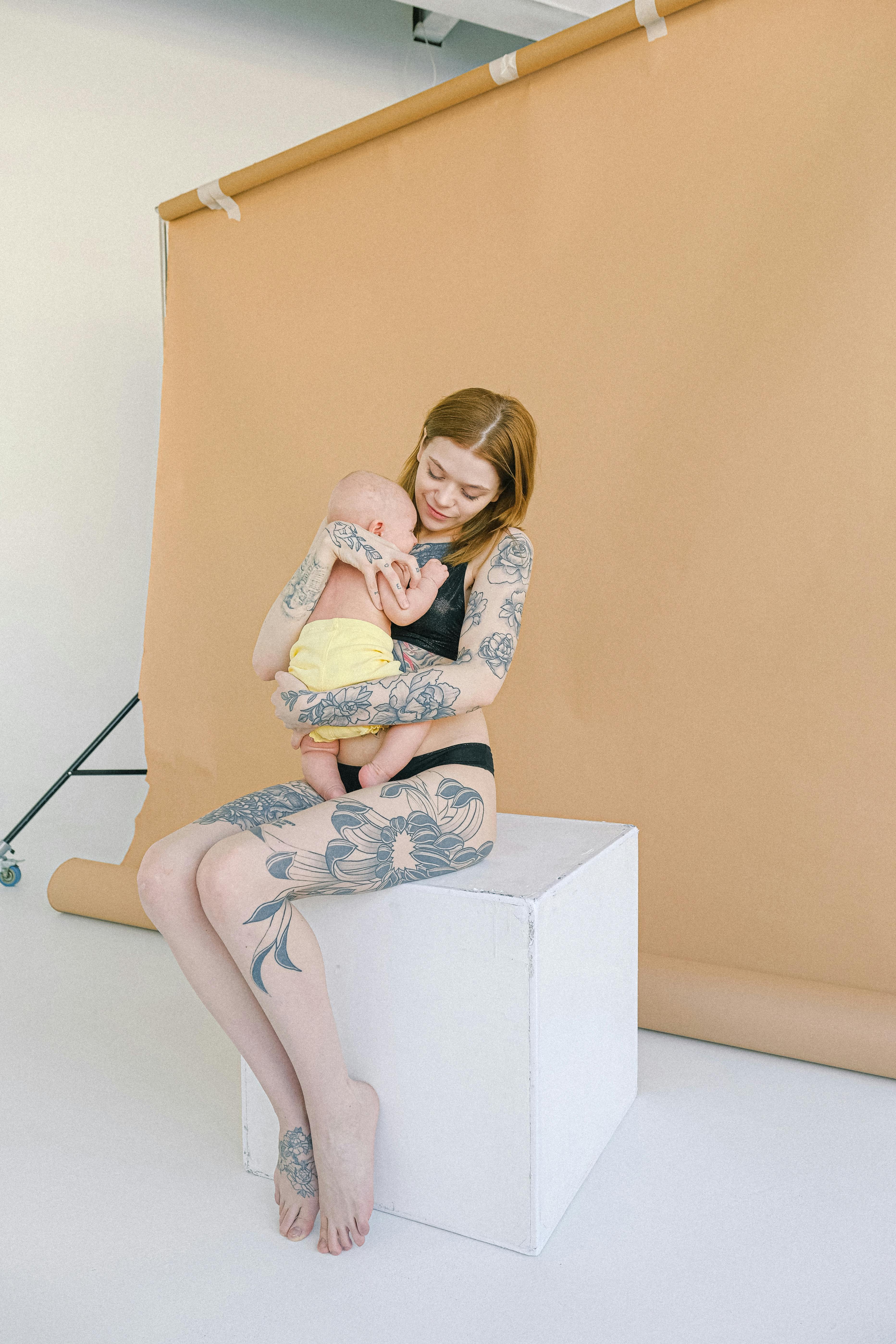 young tattooed mother in underwear embracing newborn baby in modern studio