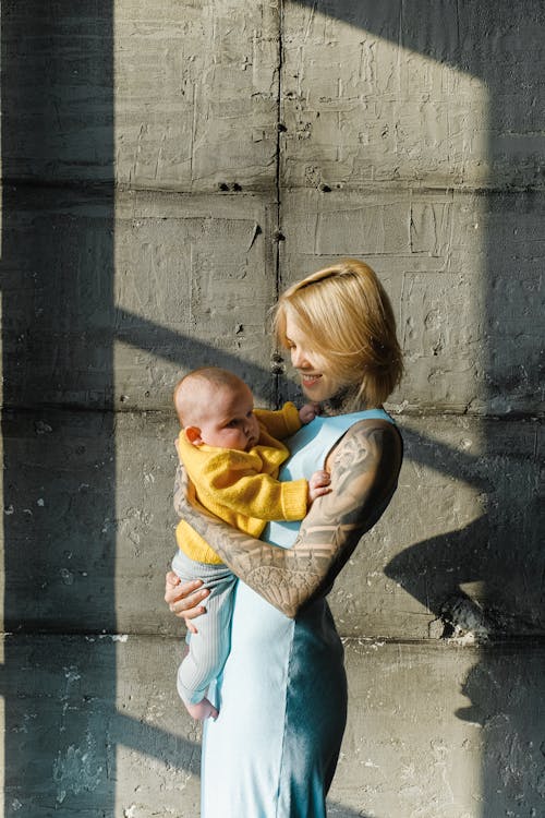 Free Side view of smiling tattooed woman in casual clothes embracing cute infant child while standing against gray concrete blocks in daytime Stock Photo