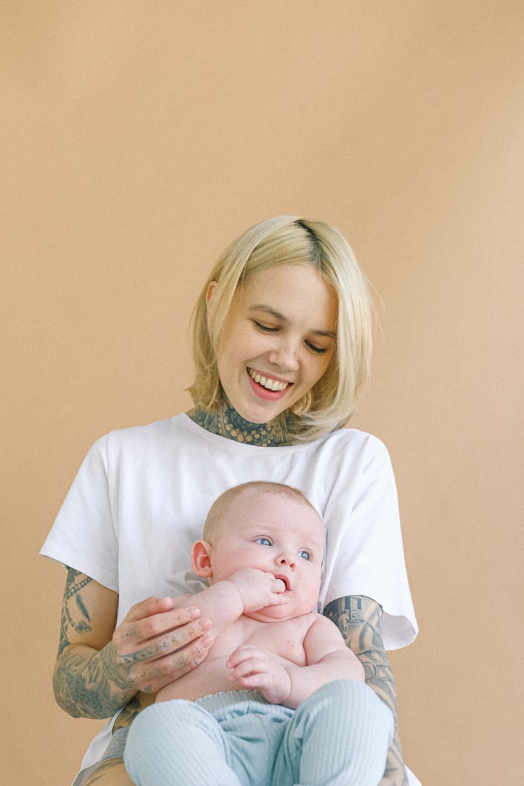 Laughing Young Mother Hugging Cute Newborn While Baby Holding Fingers In Mouth In Nursery