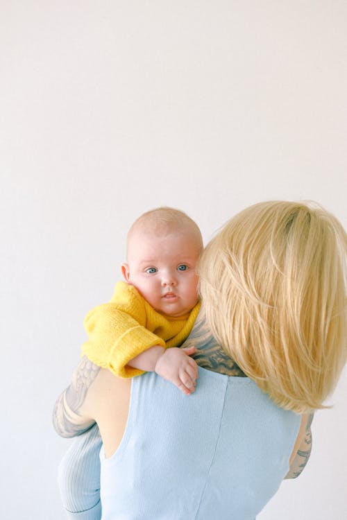무료 스튜디오에서 회색 배경에 작은 아기를 들고 문신을 한 금발 어머니 스톡 사진