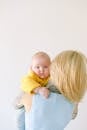 Back view of female in blue top embracing adorable newborn kid in yellow sweater while babe looking at camera in room