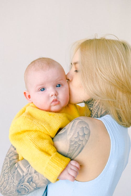 無料 部屋の腕の中で幼児の赤ちゃんにキスする愛情のある入れ墨のお母さん 写真素材