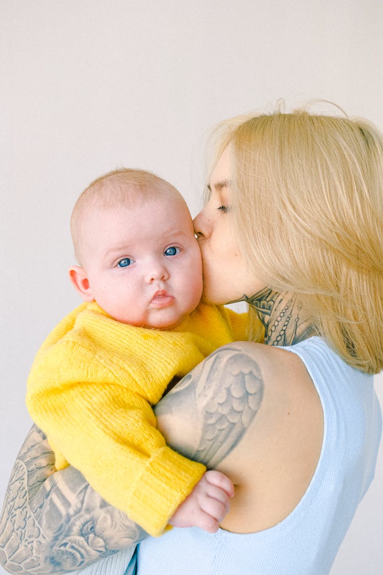 Loving Tattooed Mom Kissing Infant Baby In Arms In Room