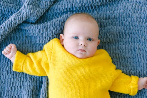 Top view of adorable baby in yellow sweater looking at camera while resting on soft blue blanket in room at daytime