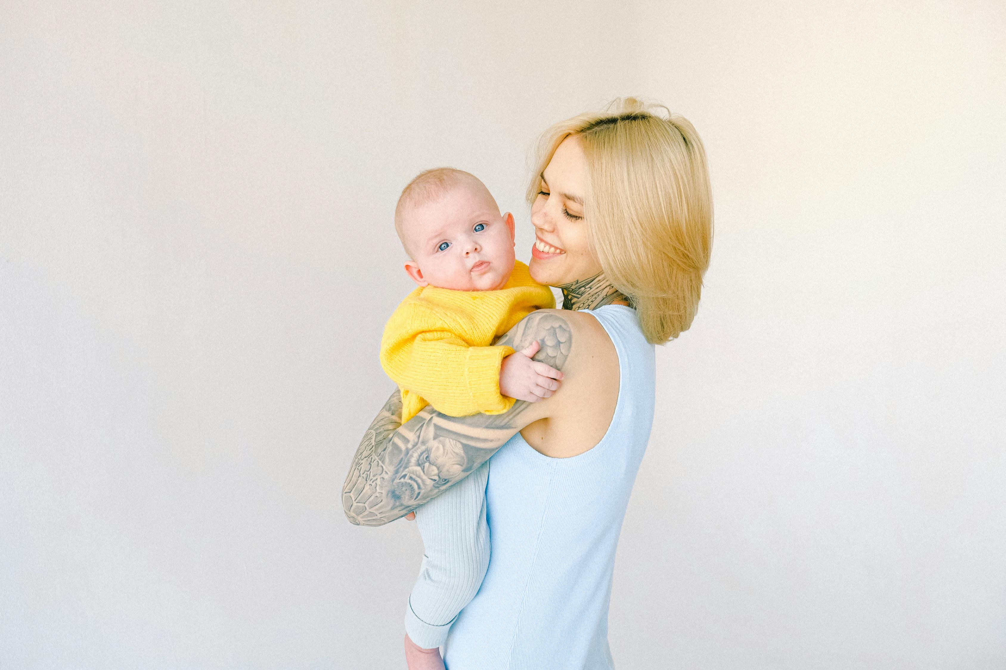 Cheerful tattooed female hugging little baby in arms against gray background