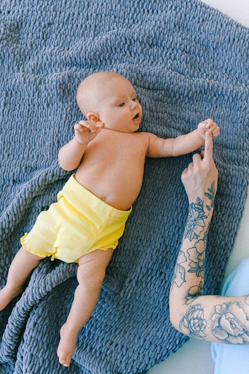 Crop tattooed female with curious infant baby lying on soft blanket in bed