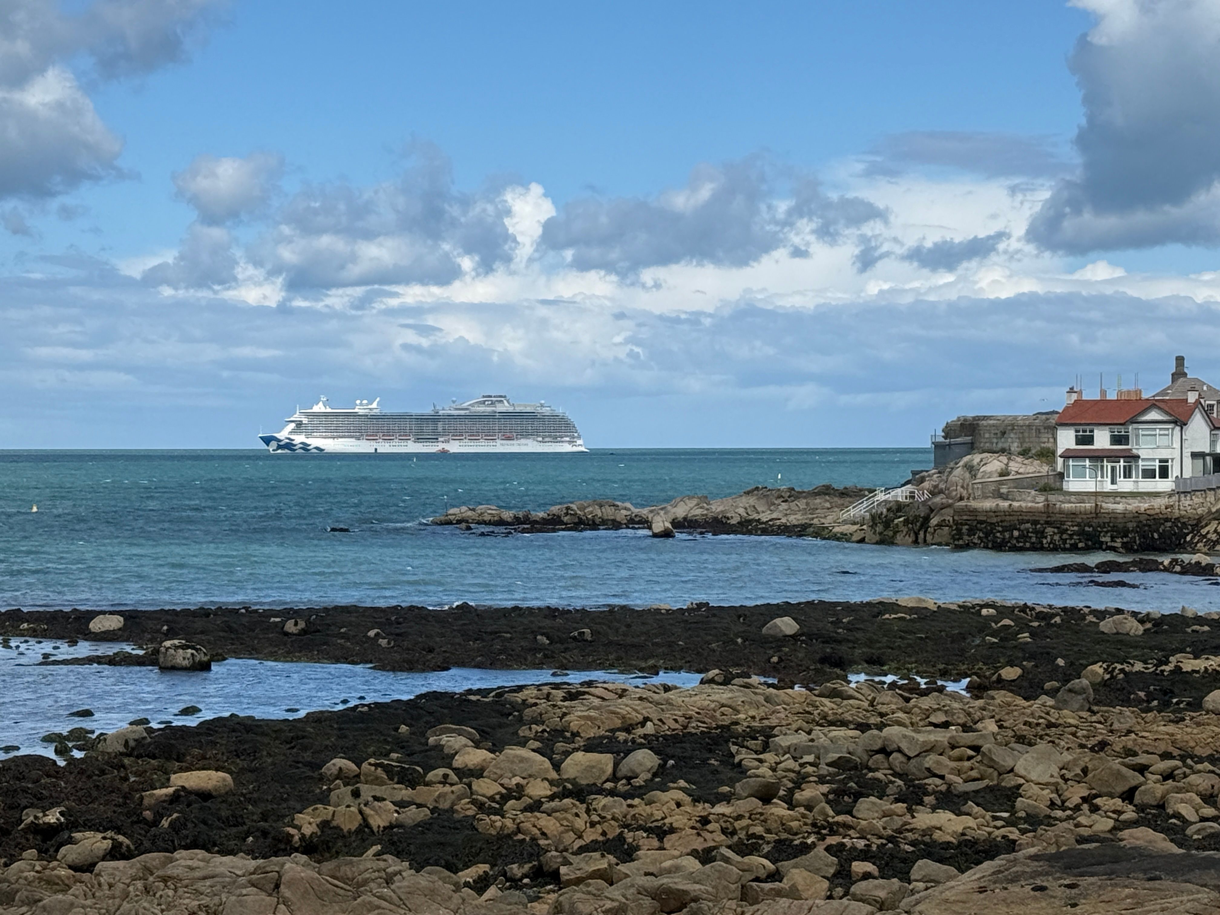 A serene coastal scene in Dalkey, Ireland, featuring a cruise ship on the horizon and rocky shores in the foreground.