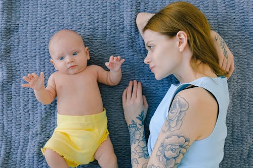 Free Top view of tattooed female in casual clothes looking at cute infant while resting together on soft blanket in room Stock Photo