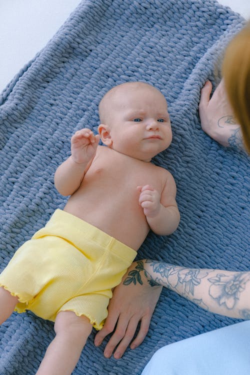 From above of adorable toddler in diaper and yellow shorts lying on soft plaid and looking at unrecognizable tattooed mom during afternoon nap