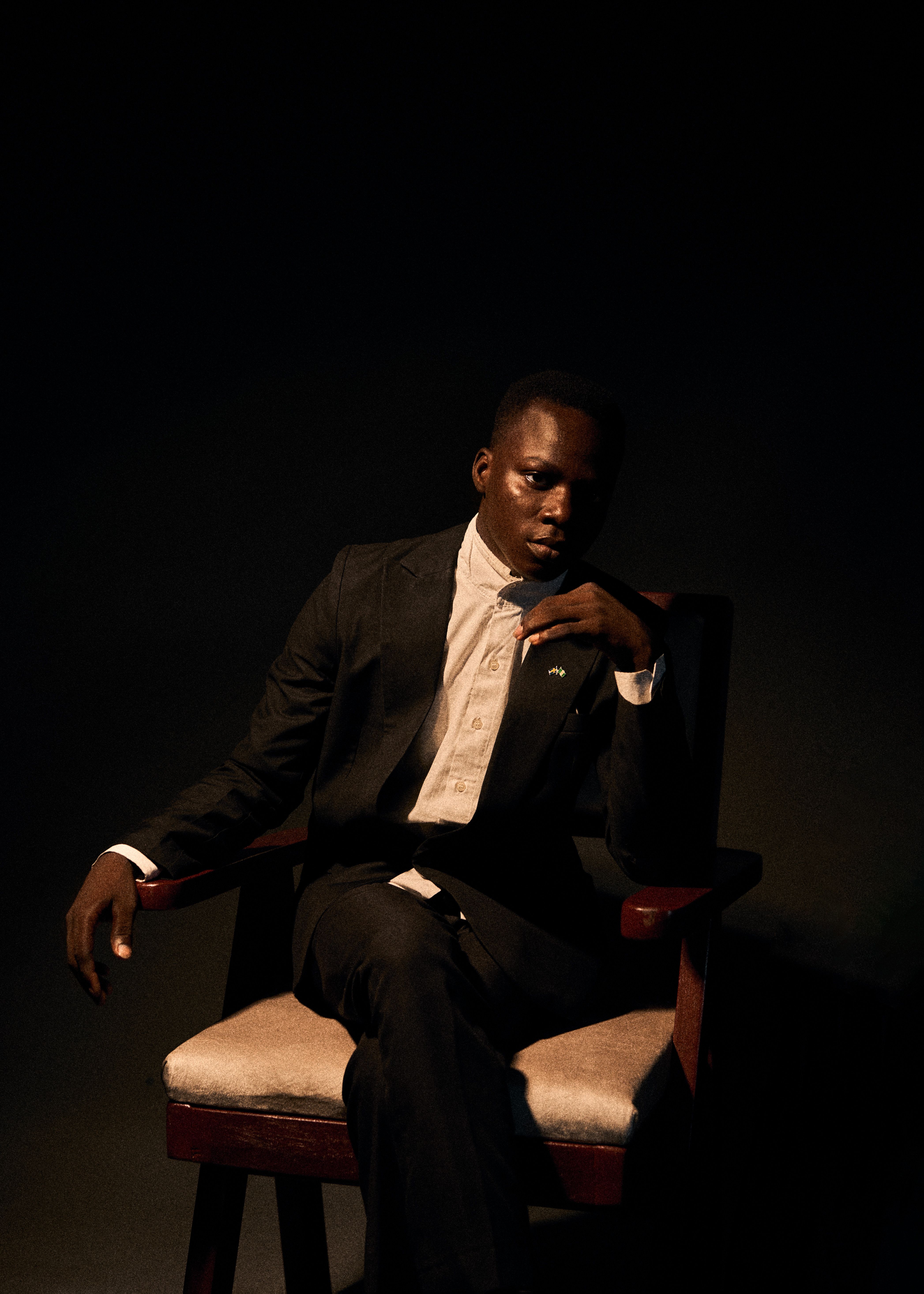 A contemplative man in a suit sits in shadowy lighting, exuding a moody atmosphere.