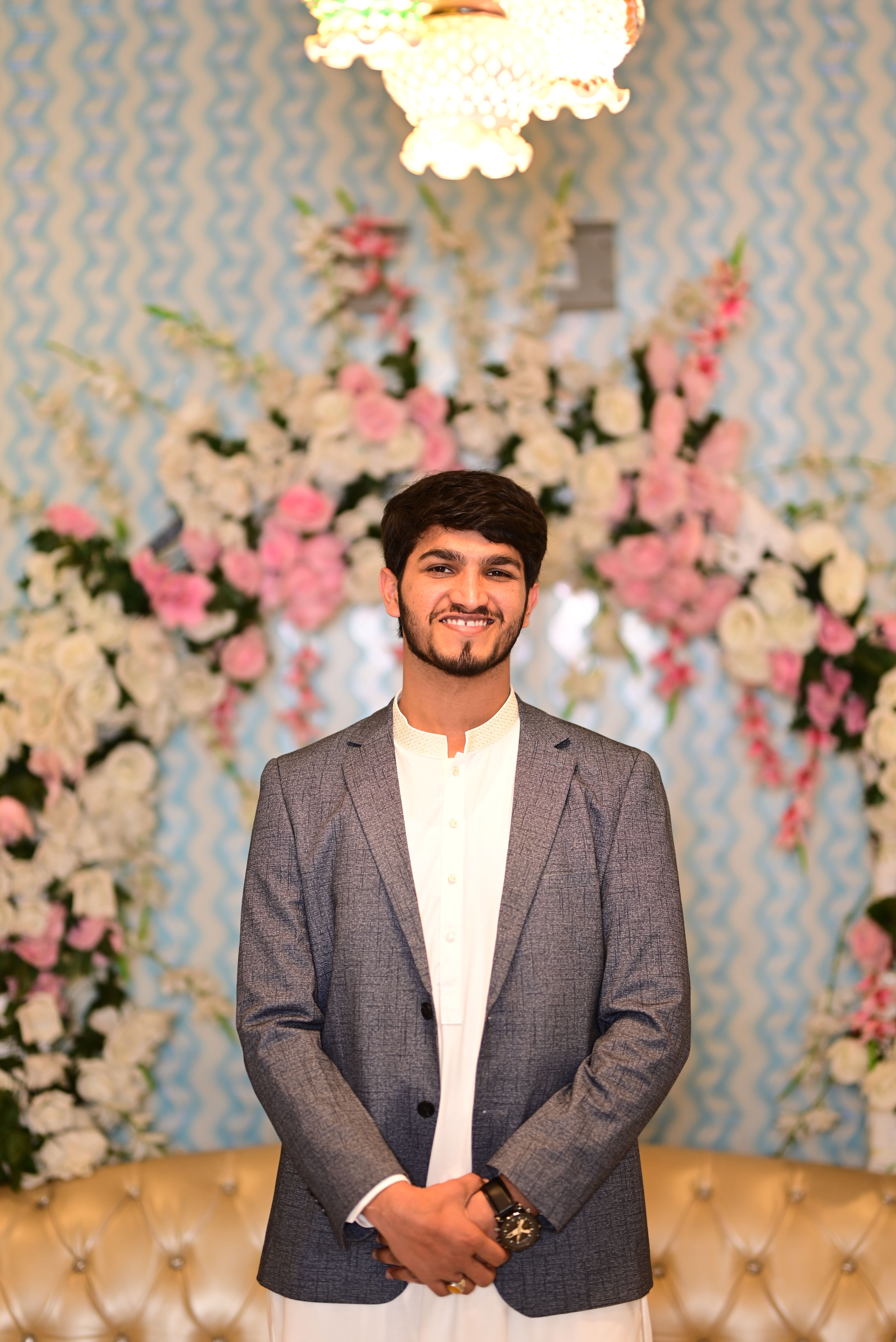 Man in traditional formal attire standing indoors with floral background.
