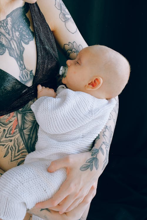 Free Crop tattooed mother holding sleeping baby with pacifier in mouth in studio Stock Photo