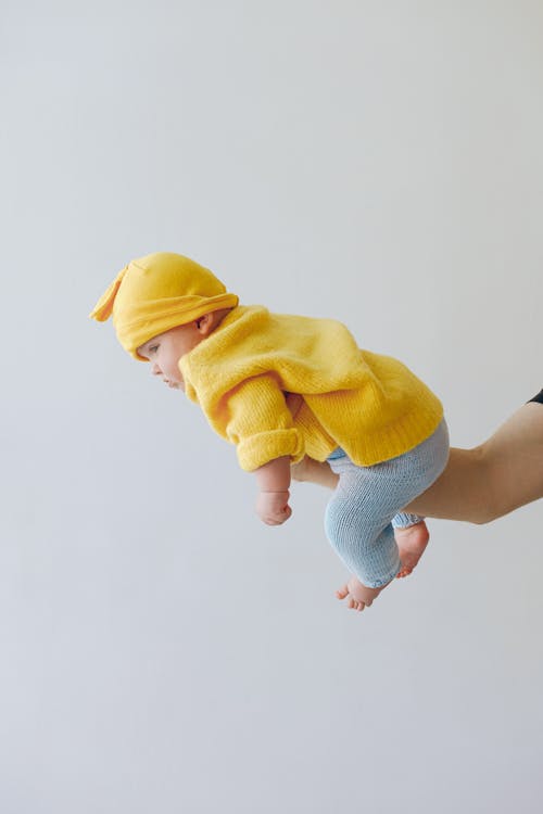 Side view full body of cute infant in warm clothes looking away while being carried by unrecognizable parent in studio