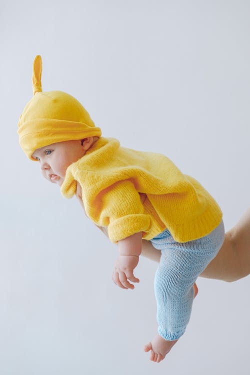 Little baby in funny hat supported by crop parent against gray background in studio