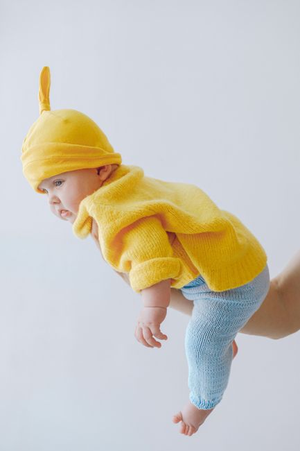 Side view full body of cute infant baby in colorful outfit barefoot hanging on outstretched arm while spending time in light room