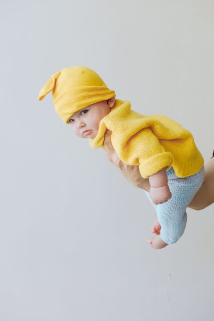 Charming baby in yellow knit outfit held in a playful flying pose indoors.