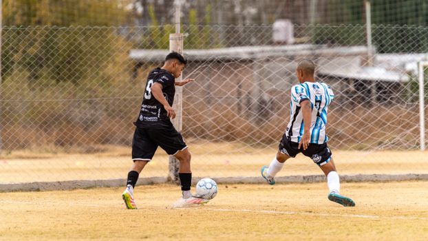 Two male soccer players in action on a grass field during a daytime match with chain-link fence backdrop.