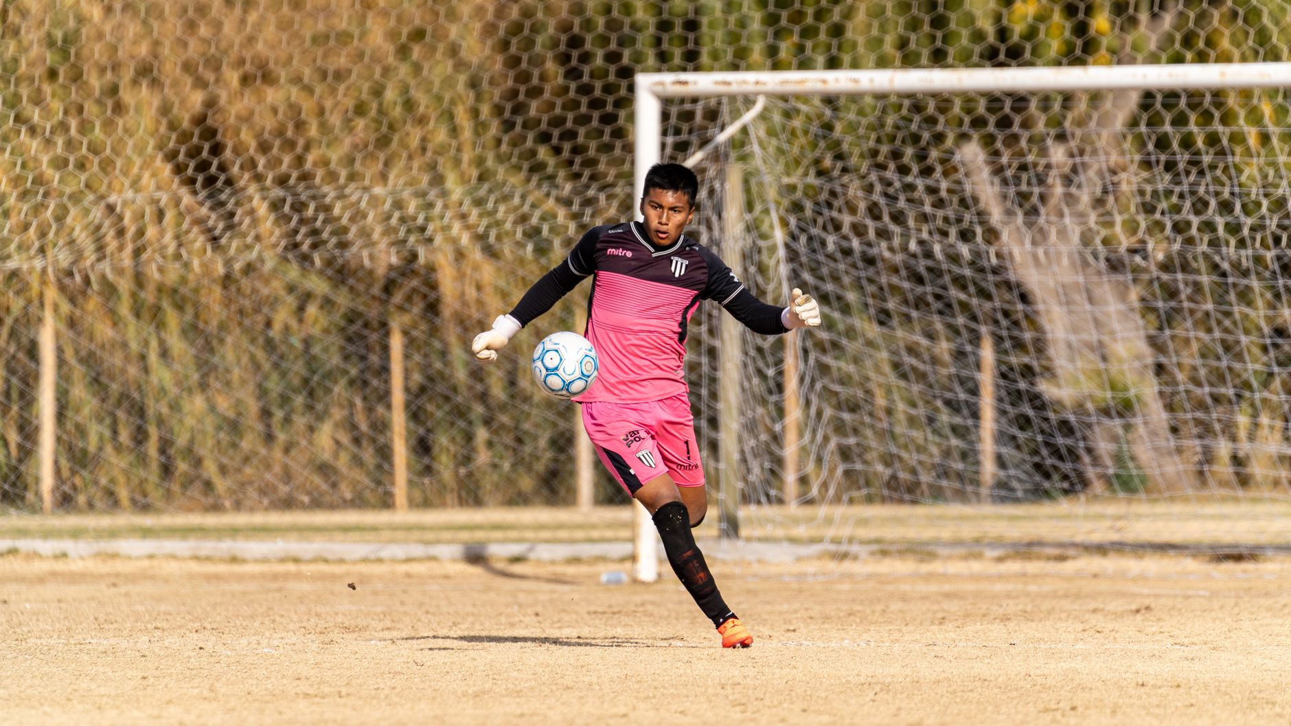 A young goalkeeper in a pink uniform kicks a soccer ball during a daytime practice on a grassy outdoor field.