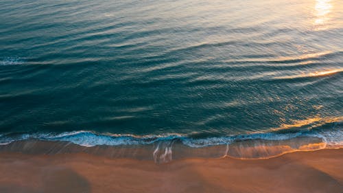 Drone picturesque view of of calm waves rolling on sandy coast and sun reflecting on turquoise water during sunset