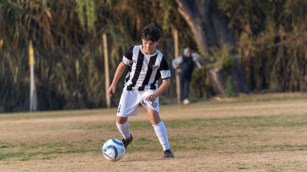Young soccer player in a black and white uniform dribbles the ball on a grassy field during a sunny day.