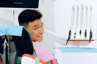 Back view of smiling ethnic man sitting in chair of dentist while preparing for treatment in modern clinic and looking over shoulder at camera