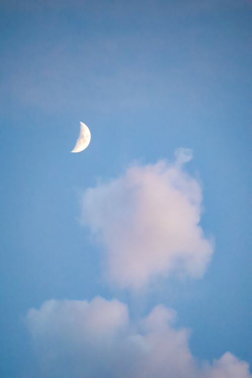 Free A serene view of the half moon and fluffy clouds set against a clear blue sky captured in daylight. Stock Photo