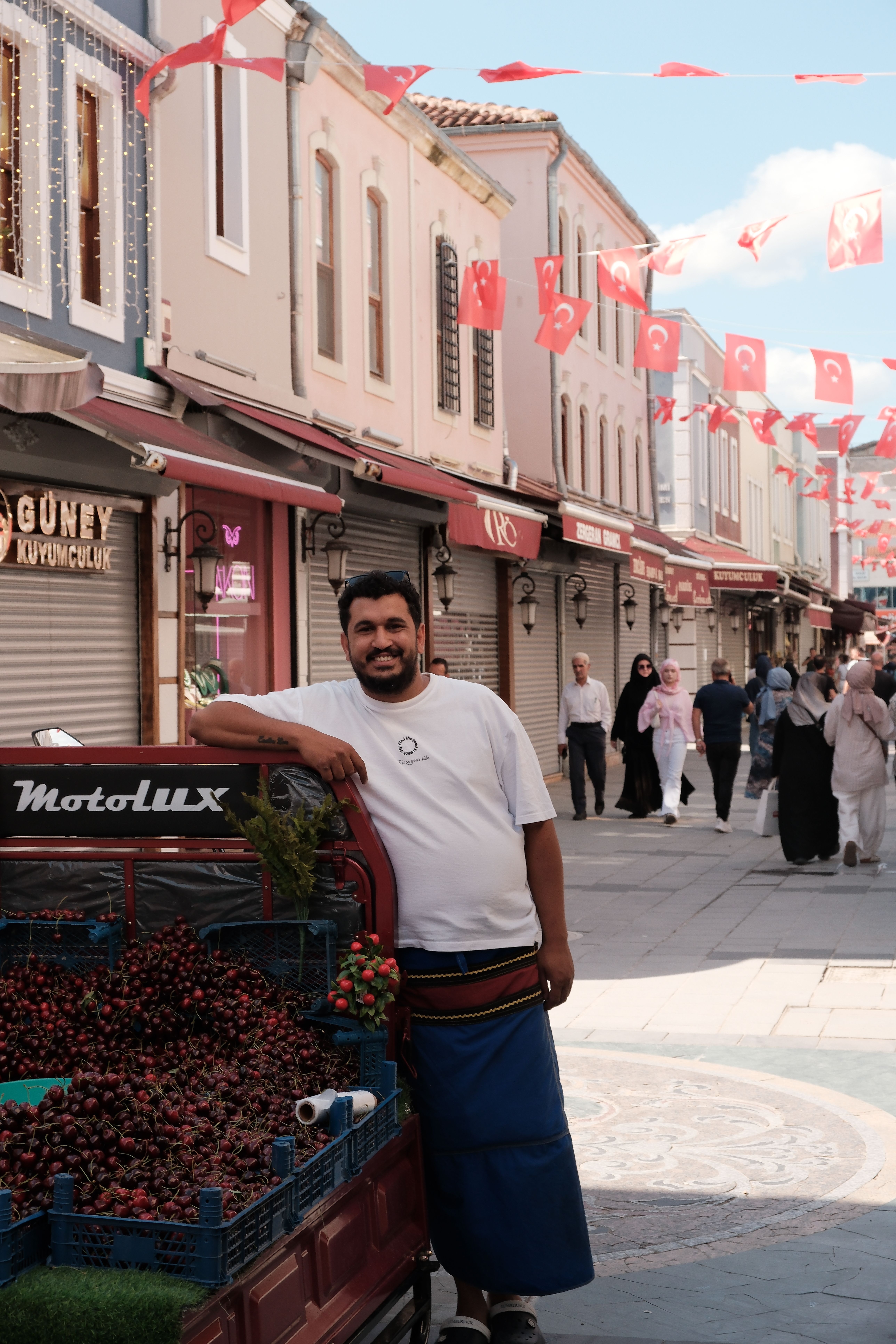 Vibrant street market in Sakarya with smiling vendor displaying fresh produce, Turkish flags adorning the scene.