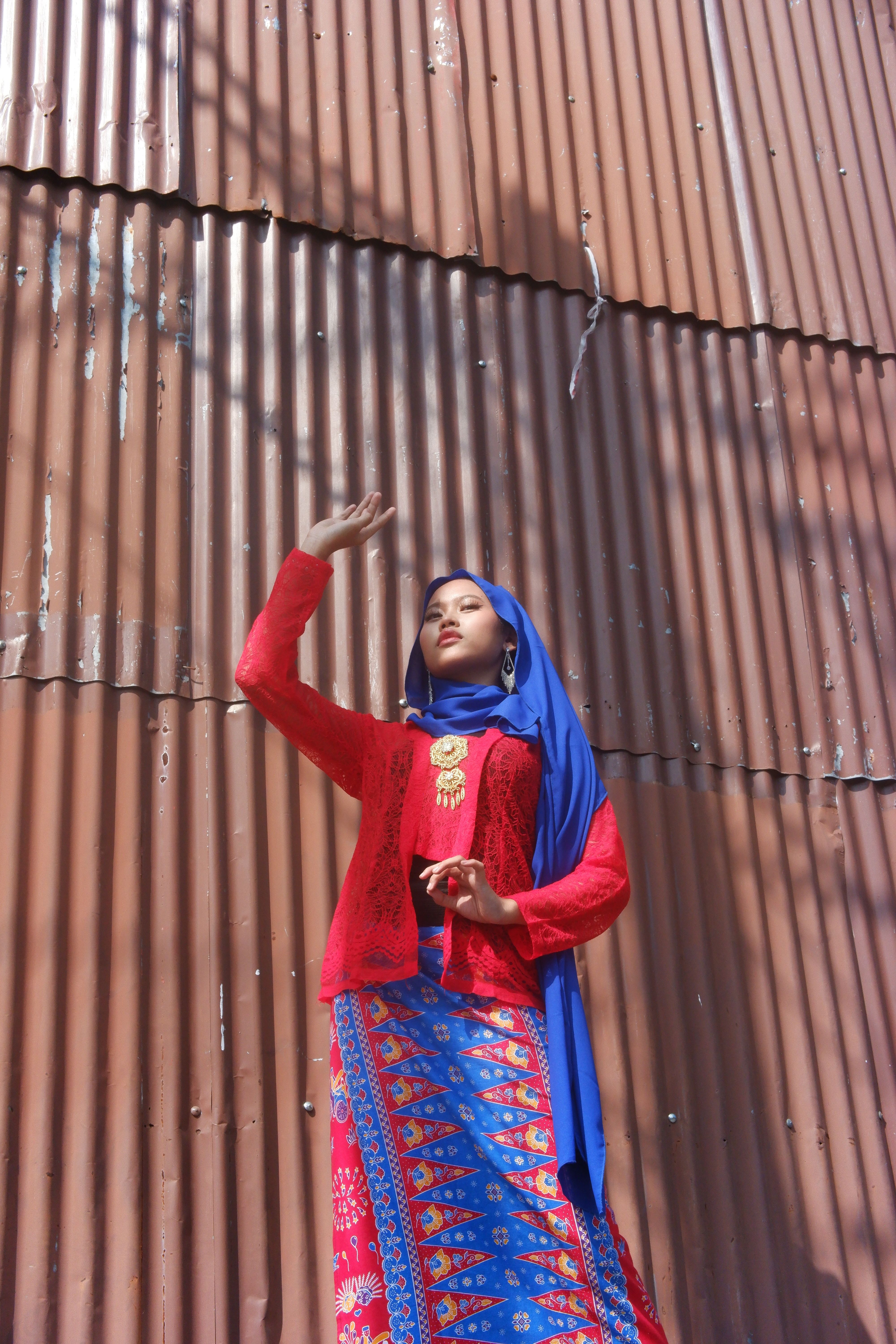 A woman in vibrant traditional clothing poses outdoors against a corrugated metal backdrop.