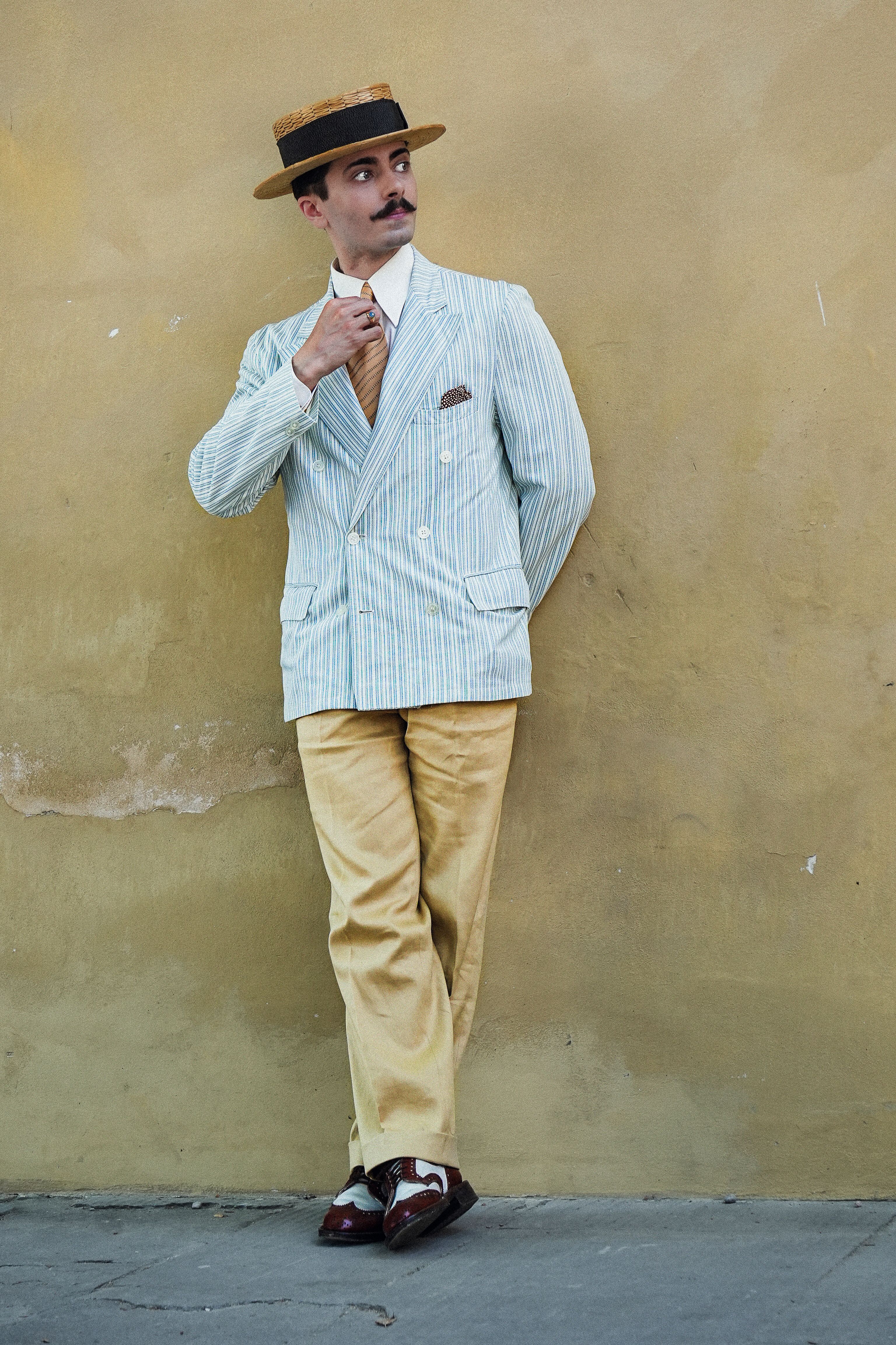 Man dressed in vintage summer attire with hat, posing against a rustic wall.