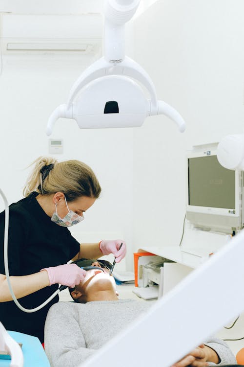 Focused woman in latex gloves and medical mask wearing black uniform holding medical instruments while treating teeth of male client in modern dental room