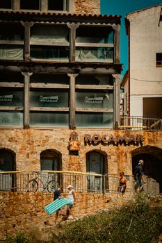 A rustic stone facade of a building in Tossa de Mar, Spain, with people walking by. Captured on a sunny day.