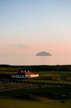 Beautiful sunset view of Ailsa Craig from a golf course featuring lush green fields and a clubhouse.