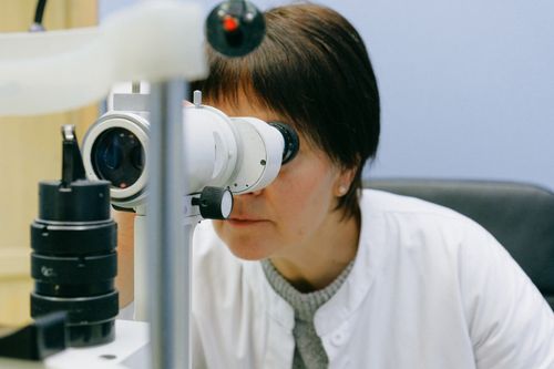 Free Anonymous female biologist in white coat observing object through big lab microscope while sitting on grey chair and leaning forward in light workplace Stock Photo